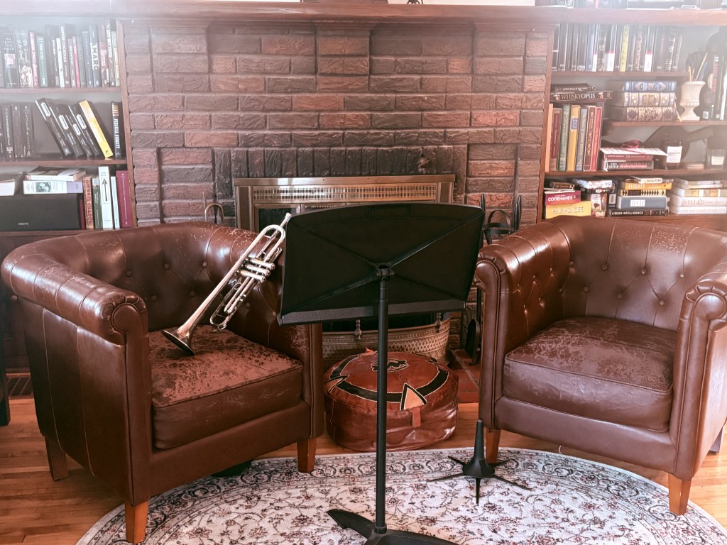 Two cozy chairs and a music stand in front of a fireplace. One chair has a trumpet resting on it.